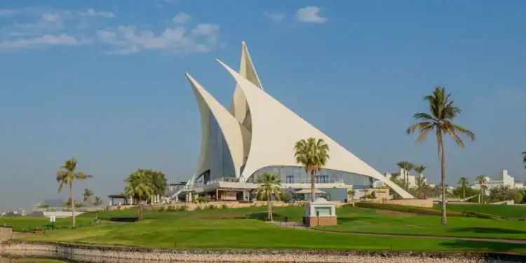 Dubai Creek Hosts 2025 International Pairs Golf Event The iconic sail-shaped clubhouse of Dubai Creek Golf & Yacht Club on a sunny day, with lush green fairways, a water hazard in the foreground, and palm trees under a blue sky.