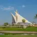 Dubai Creek Hosts 2025 International Pairs Golf Event The iconic sail-shaped clubhouse of Dubai Creek Golf & Yacht Club on a sunny day, with lush green fairways, a water hazard in the foreground, and palm trees under a blue sky.